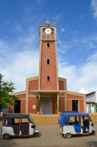 The clock tower stands prominently in Villanueva, Santander, with colorful tuk-tuks parked in front against a bright blue sky.