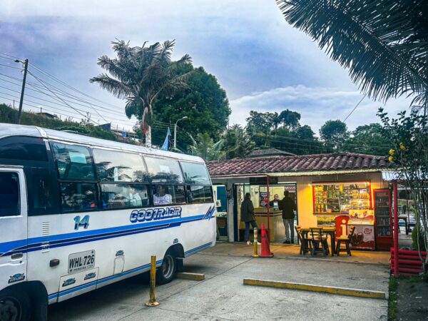 Colorful bus station in Salento, Quindío, Colombia, featuring a white bus, small shop, and vibrant greenery.