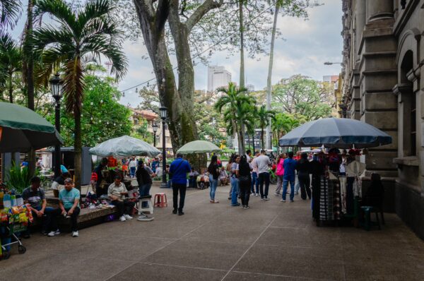 Plazuela San Ignacio in Medellín with people gathered under trees, market umbrellas, and historic buildings along the square