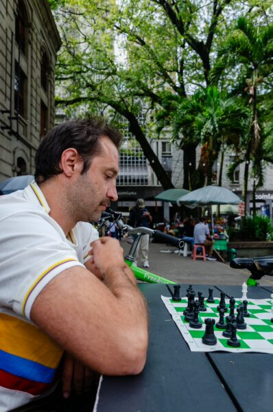 A man concentrated on a chess game in a lively outdoor plaza surrounded by tropical trees in San Ignacio, Medellín.