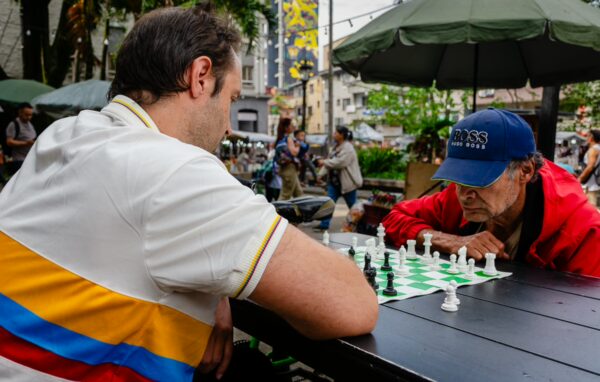 Two men play chess at an outdoor table in Plazuela San Ignacio, Medellín, with umbrellas and pedestrians in the background