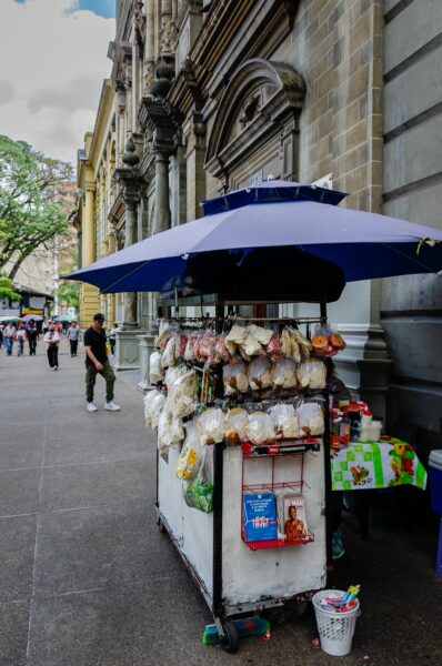 Vibrant food cart under a blue umbrella, with colorful snacks, set against the historic architecture in San Ignacio, Medellín.