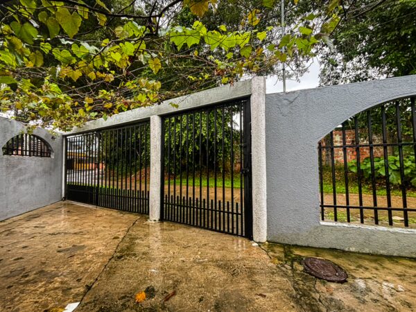 Black metal gates framed by green foliage in Leticia, Amazonas, Colombia, with a wet stone pathway and greenery in the background.