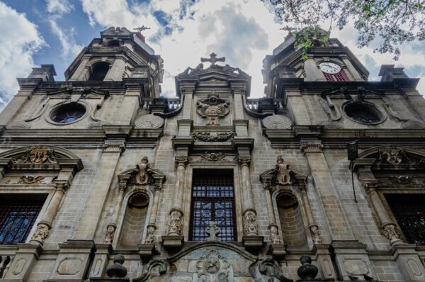 The ornate facade of Iglesia de San Ignacio in Medellín, featuring intricate carvings and tall, decorative towers against a cloudy sky.