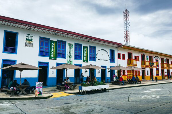 Colorful colonial buildings line Plaza Bolivar, with vibrant blue and yellow accents, outdoor seating, and people enjoying the lively atmosphere in Salento, Quindio, Colombia.