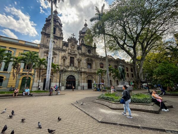 Plaza in Medellín features an ornate church with towering spires, surrounded by palm trees and people enjoying the sunny day.