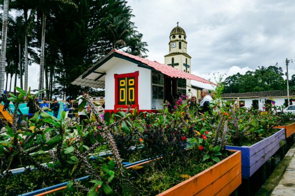 Colorful plants in vibrant flower beds surround a charming house with yellow doors and a clock tower in Plaza Bolívar, Salento, Quindío.