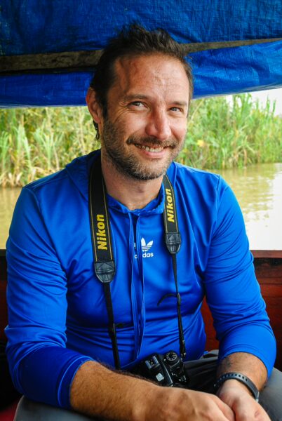 Smiling man in a blue shirt and camera around his neck, with a lush green riverbank in Gamboa Nature Reserve, Loreto, Peru.