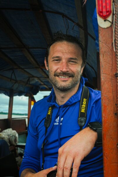 A smiling traveler wearing a blue shirt and camera strap, enjoying a boat ride through Gamboa Nature Reserve, Loreto, Peru.