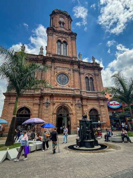 Iglesia San José in Medellín features stunning brick architecture, surrounded by vibrant umbrellas and palm trees against a blue sky.