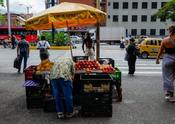 Colorful fruit stand bustling with people in Medellín, Colombia, featuring fresh apples and bananas under a yellow canopy.