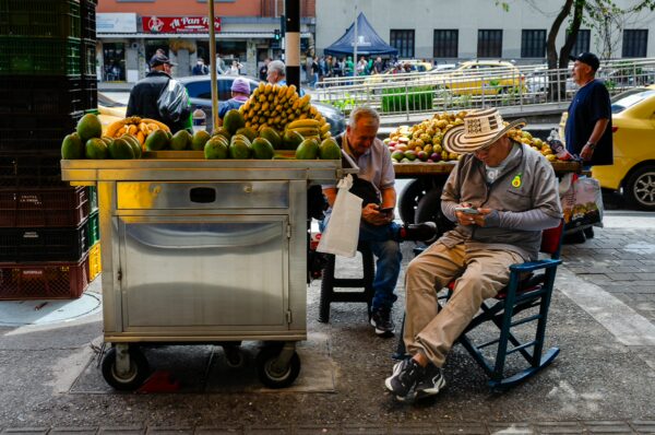 Vibrant street scene in Medellín, featuring two men by a fruit cart full of tropical fruits and colorful urban surroundings.