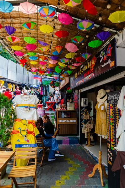Vibrant umbrellas hang above a colorful alleyway in Comuna 13, Medellín, showcasing local art, clothing, and lively street life.