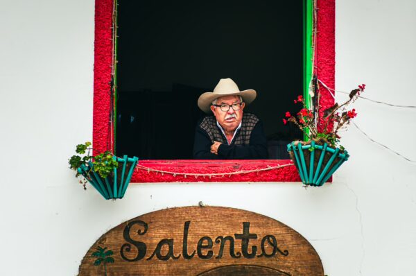 An elderly man with glasses and a cowboy hat leans out of a colorful window in Salento, Quindio, surrounded by potted flowers.