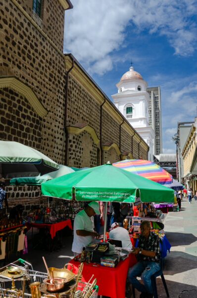 Street market beside the Basilica of Our Lady of Candelaria in central Medellín, with vendors under colorful umbrellas and a white church tower behind