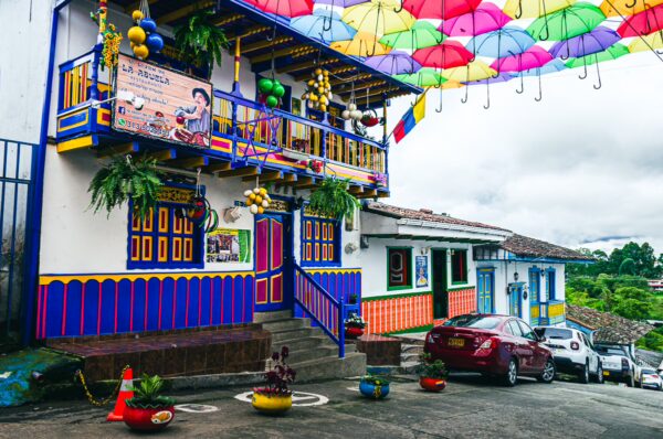 Colorful umbrellas hang above Calle Real in Salento, Quindío, highlighting vibrant buildings adorned with plants and decorations.
