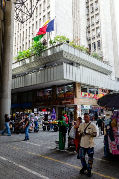Street market beneath an elevated structure in central Medellín, with vendors, shoppers, and flags above the plaza