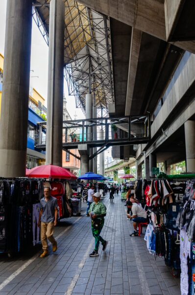 Street market under the elevated San Antonio Metro tracks in central Medellín, with clothing stalls and pedestrians in the shade
