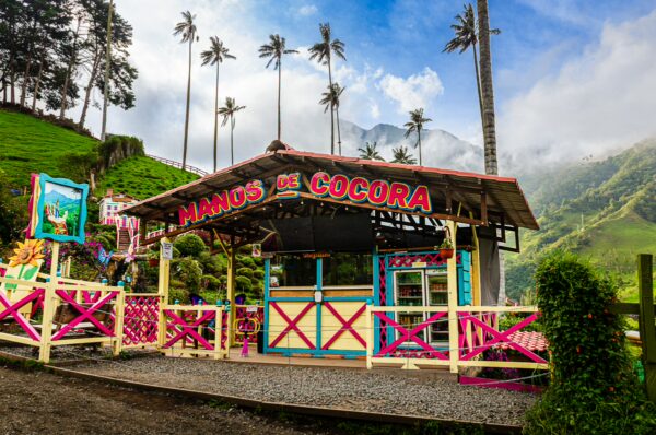 Brightly colored kiosk named "Manos del Cocora" nestled among lush green hills and towering wax palms in Cocora Valley, Quindío, Colombia.