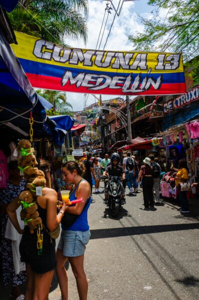 Vibrant streets of Comuna 13 in Medellín, Colombia, bustling with vendors, colorful banners, and lively crowds.