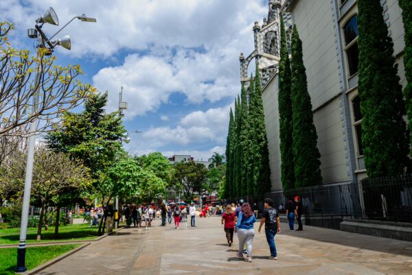 Visitors stroll through Plaza Botero in Medellín, surrounded by lush greenery and vibrant sculptures, under a partly cloudy sky.