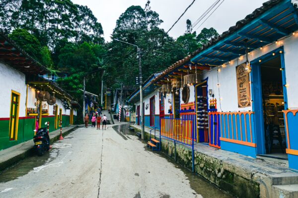 Calle Real in Salento, Quindío, features vibrant houses, charming shops, and lush green hills under a cloudy sky.