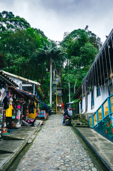 Colorful shops line the cobblestone Calle Real, leading towards lush greenery and a path in Salento, Quindío, Colombia.