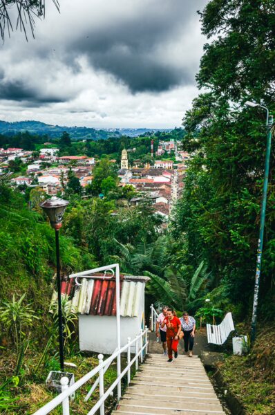 Colorful houses dot the hillside as visitors descend steps toward Salento's lush valley, under a dramatic, cloudy sky.