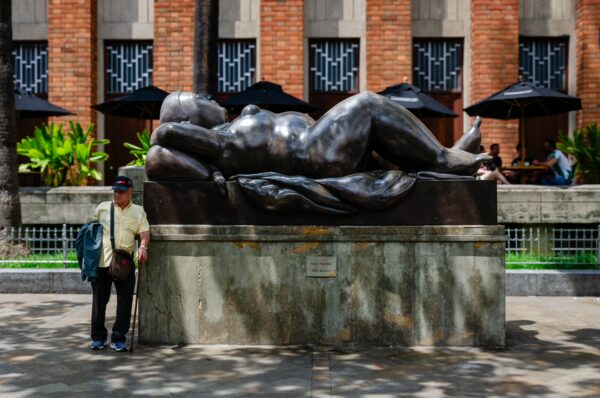 A visitor leans against a large bronze statue in Plaza Botero, surrounded by lush greenery and shaded seating areas.