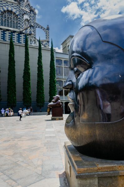 Large sculptures dominate Plaza Botero in Medellín, surrounded by lush greenery and vibrant architecture under a clear blue sky.