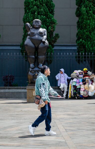 A man walks past a large, humorous sculpture in Plaza Botero, surrounded by trees and vendors displaying colorful souvenirs.