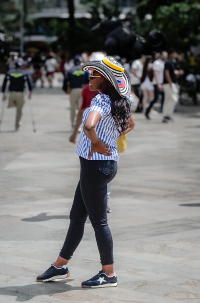 A woman in a striped shirt and colorful hat poses confidently in Plaza Botero, surrounded by bustling crowds in Medellín, Colombia.