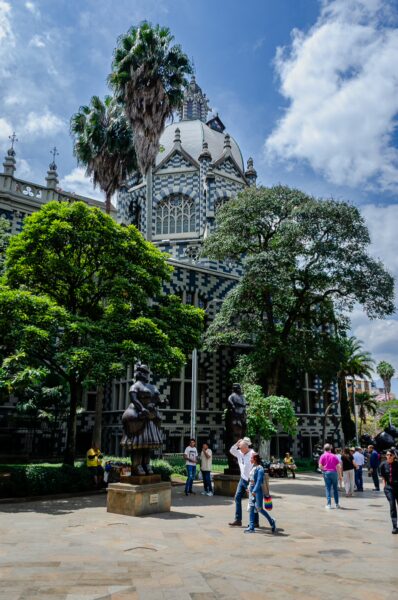 Visitors explore Plaza Botero in Medellín, surrounded by lush greenery and striking architecture under a bright blue sky.