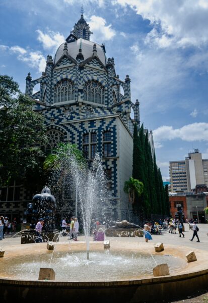 A decorative fountain splashes water in front of a colorful building, framed by lush greenery and a bright blue sky in Medellín.