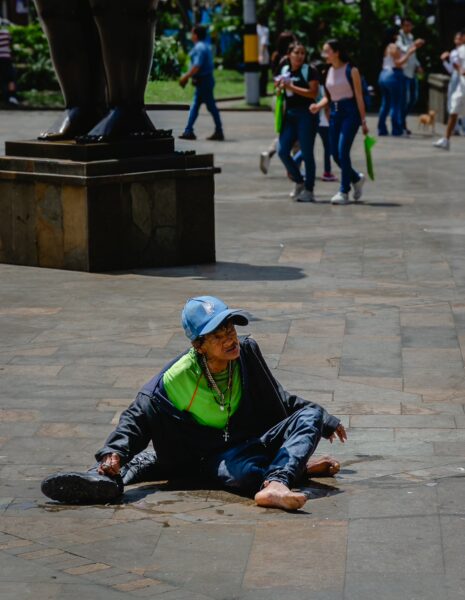A person sits on the pavement in Plaza Botero, surrounded by visitors and green trees in Medellin, Antioquia.