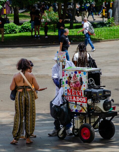 A woman in patterned trousers engages with a colorful ice cream cart in Plaza Botero, surrounded by green parks and visitors.