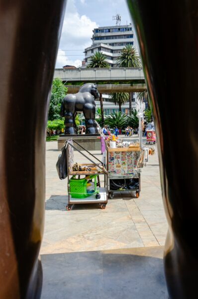 A view through a sculpture at Plaza Botero, showcasing a large gorilla statue and a colorful vendor cart in Medellín.