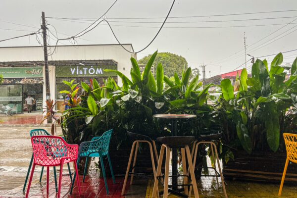 Colorful chairs surround a wooden table under rain in Leticia, Amazonas, Colombia, with lush green plants providing a tropical vibe.
