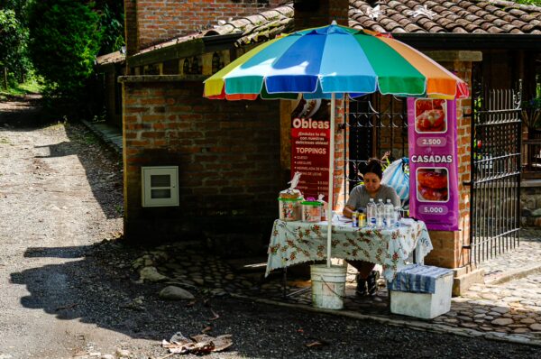 Colorful umbrella shading a vendor with snacks at Santa Rita Waterfalls in Salento, Quindío, Colombia. Cobblestone path nearby.