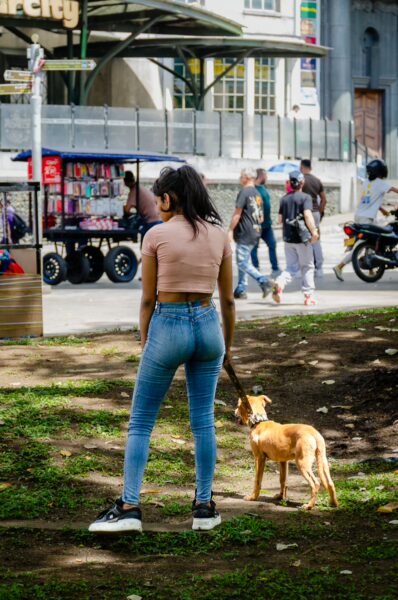 A woman in jeans stands with a dog in Plaza Botero, while pedestrians and a colorful vendor cart are seen in the background.