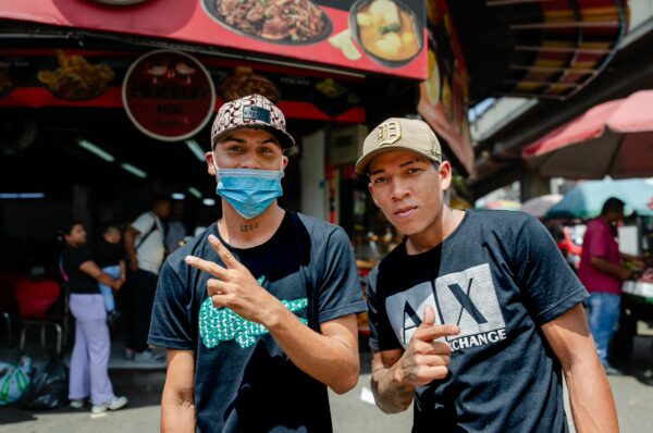 Two young men posing on a busy Medellín street, one wearing a face mask and both making hand gestures