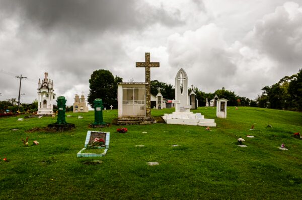 Cemetery in Salento, Quindio, Colombia, featuring ornate white tombstones, a large wooden cross, and green grass under a cloudy sky.
