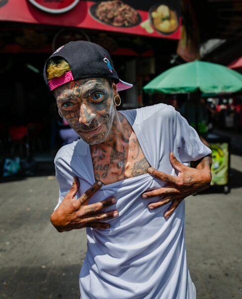 A tattooed man poses playfully in a bustling Medellín street, colorful food stalls and green umbrellas visible in the background.