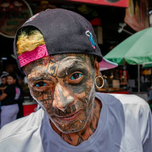 Close-up portrait of a tattooed man wearing a black cap with eyes tattooed blue and facial piercings in a Medellín street setting