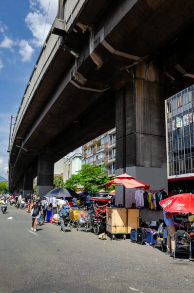Vibrant street scene in Medellín, Colombia, featuring market stalls, colorful umbrellas, and the elevated Metro line overhead.