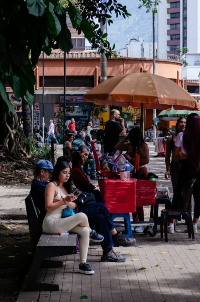 People sitting and chatting at a shaded plaza in central Medellín, with street vendors and trees around Parque de Bolívar
