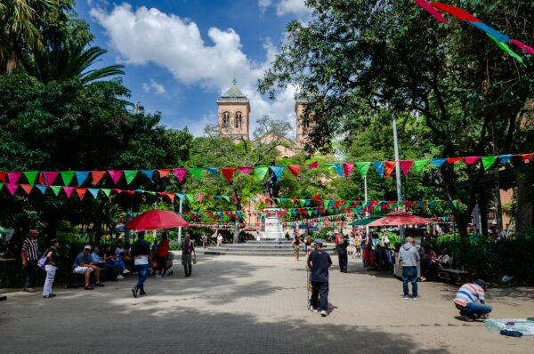 Vibrant decoration flags hang above a lively Parque Bolívar in Medellín, with visitors enjoying the lush greenery and sunny skies.