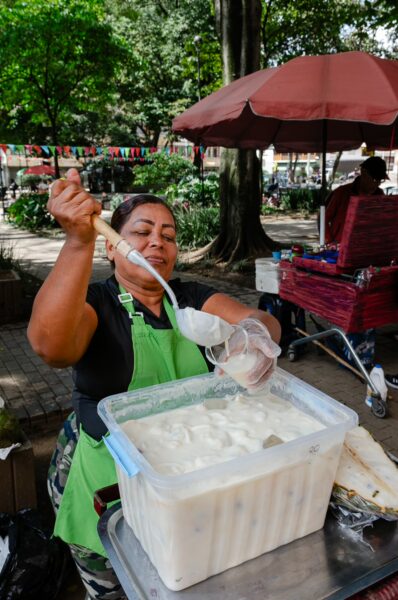 Street vendor serving a creamy guanabana juice from a large container in a shaded Medellín park