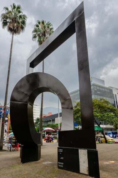 A modern black sculpture frames the sky, surrounded by palm trees and colorful market stalls in Parque San Antonio, Medellín.