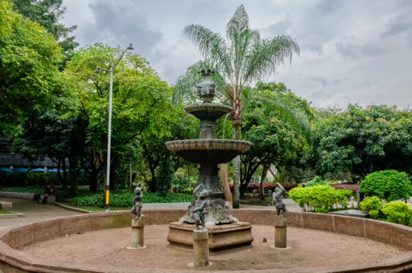 Ornate tiered fountain in Parque de San Antonio, Medellín, surrounded by trees and a cloudy sky
